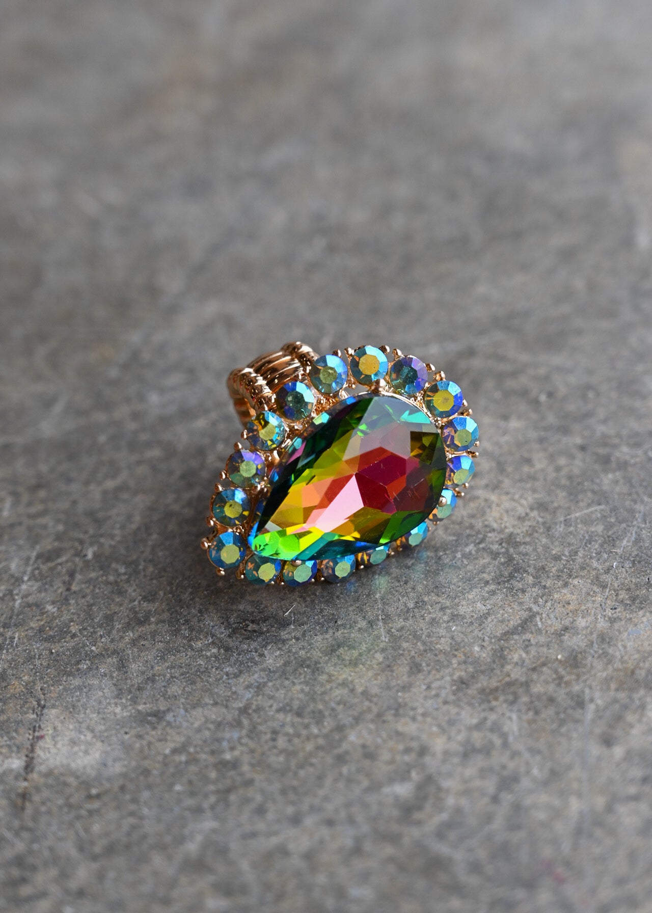 A teardrop-shaped iridescent gemstone ring with a gold-toned setting and small clear accent stones, resting on a textured gray stone surface