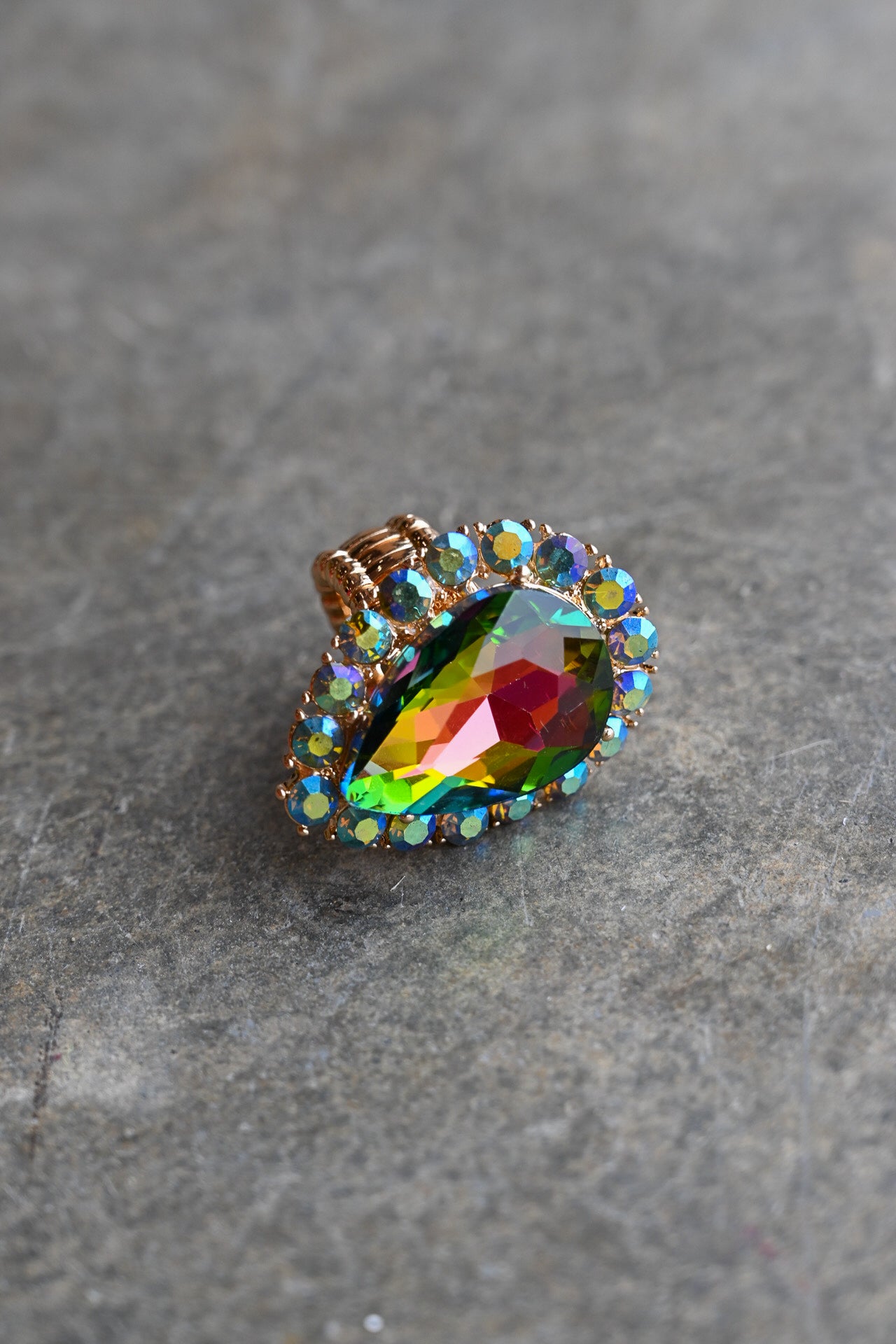 A teardrop-shaped iridescent gemstone ring with a gold-toned setting and small clear accent stones, resting on a textured gray stone surface