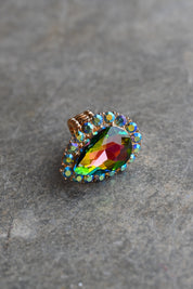 A teardrop-shaped iridescent gemstone ring with a gold-toned setting and small clear accent stones, resting on a textured gray stone surface