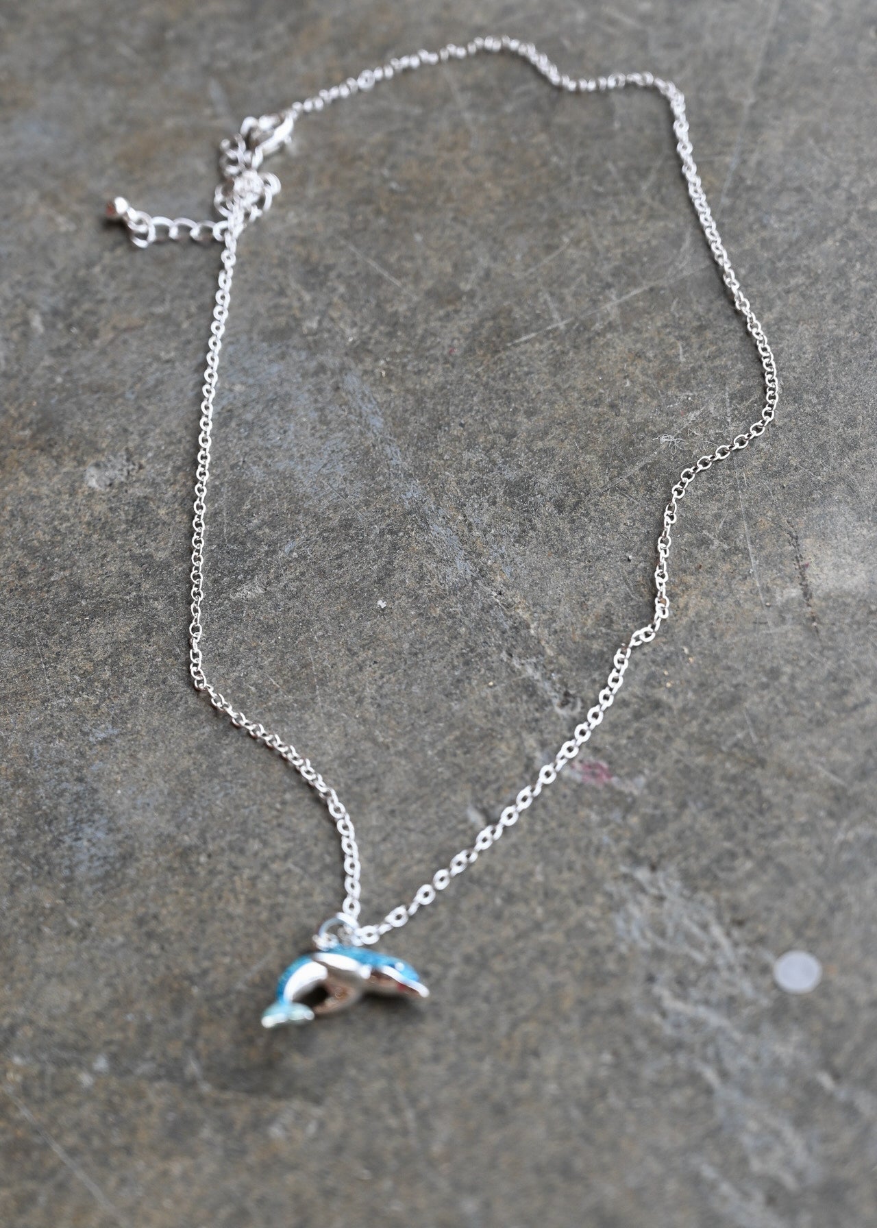 A silver chain necklace with a small dolphin pendant featuring light blue and white enamel details, displayed on a textured gray stone surface