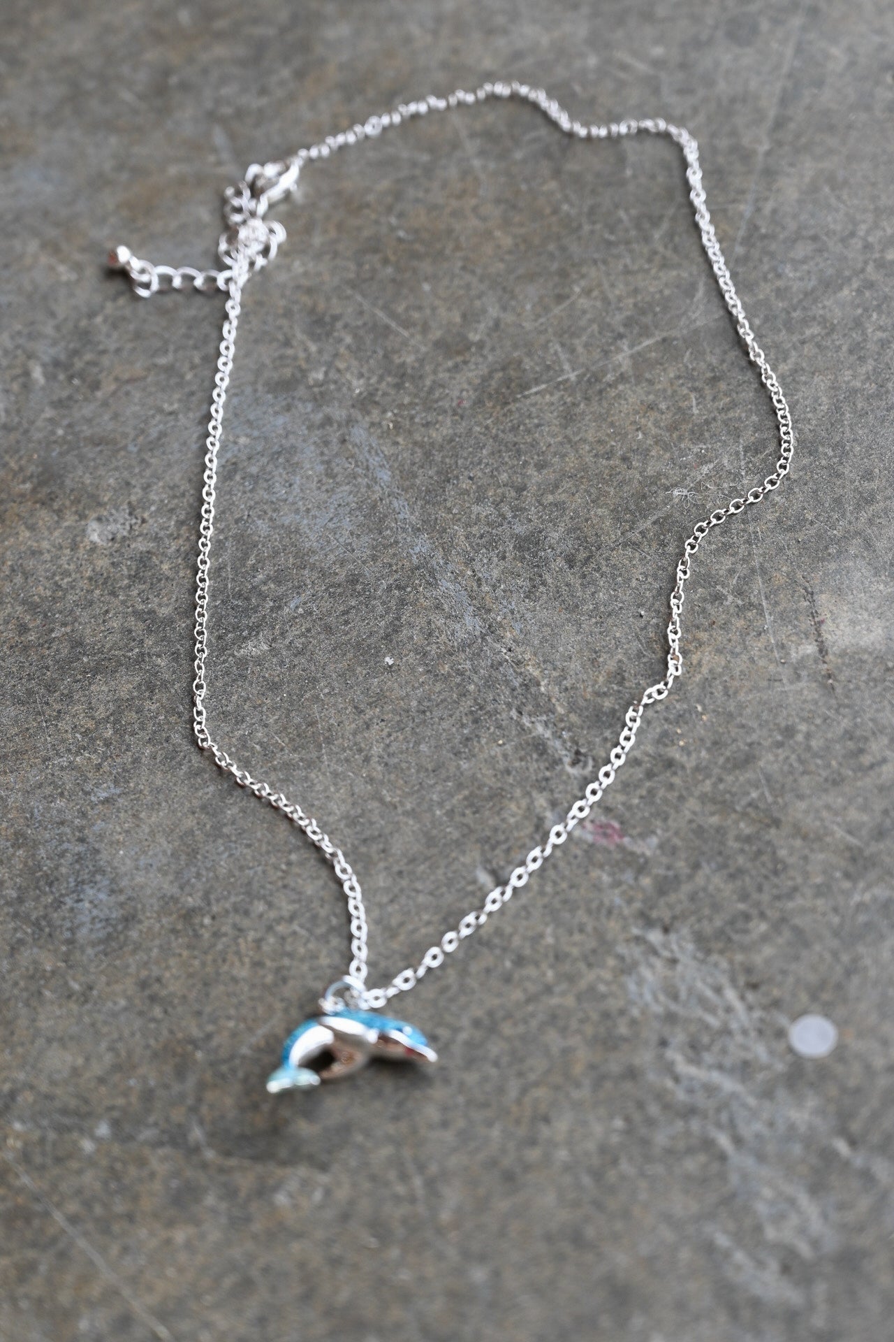 A silver chain necklace with a small dolphin pendant featuring light blue and white enamel details, displayed on a textured gray stone surface