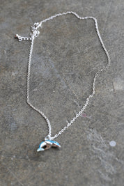 A silver chain necklace with a small dolphin pendant featuring light blue and white enamel details, displayed on a textured gray stone surface