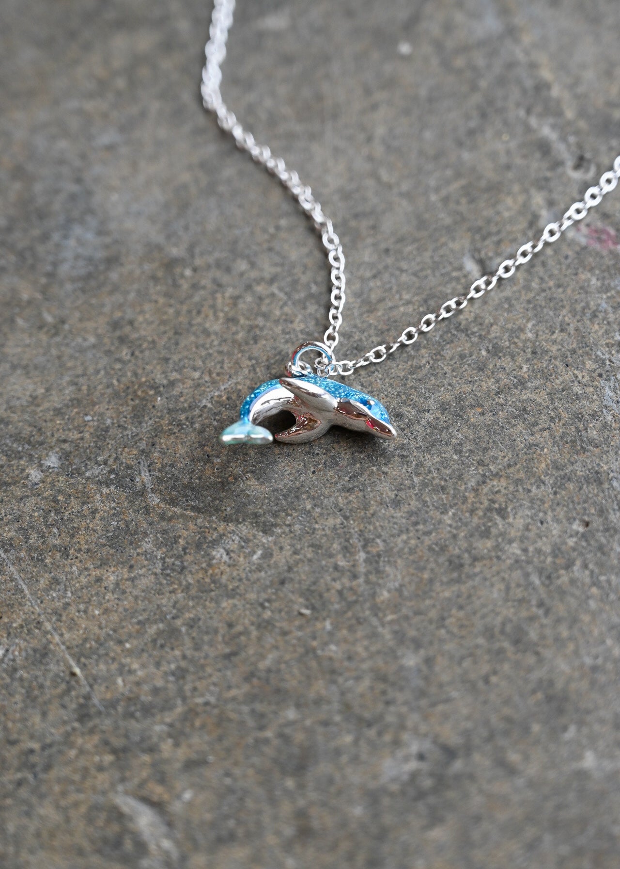 A silver chain necklace with a small dolphin-shaped pendant featuring blue and white enamel detailing, resting on a textured gray stone surface