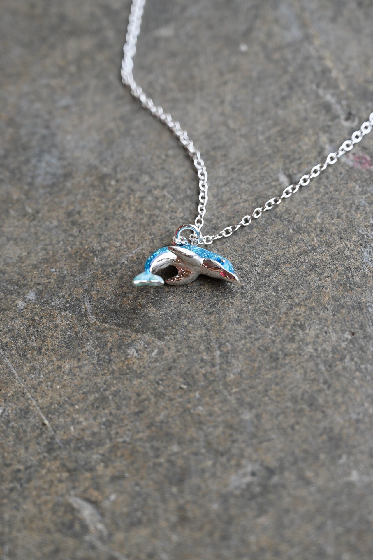 A silver chain necklace with a small dolphin-shaped pendant featuring blue and white enamel detailing, resting on a textured gray stone surface