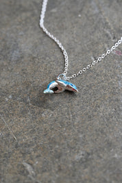 A silver chain necklace with a small dolphin-shaped pendant featuring blue and white enamel detailing, resting on a textured gray stone surface