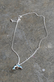 A delicate silver chain necklace with a small dolphin pendant resting on a textured gray stone surface
