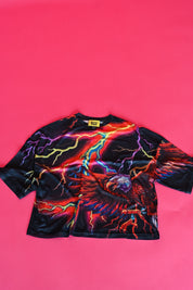 A black short-sleeve crop top featuring a vibrant, multicolored graphic print of an eagle and lightning bolts, laid flat on a solid hot pink background