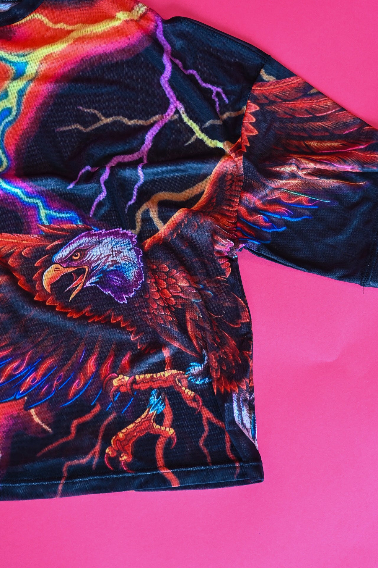 A close-up of a black short-sleeve t-shirt featuring a vibrant, colorful graphic print of an eagle with lightning bolts, laid flat on a bright pink background