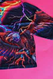 A close-up of a black short-sleeve t-shirt featuring a vibrant, colorful graphic print of an eagle with lightning bolts, laid flat on a bright pink background