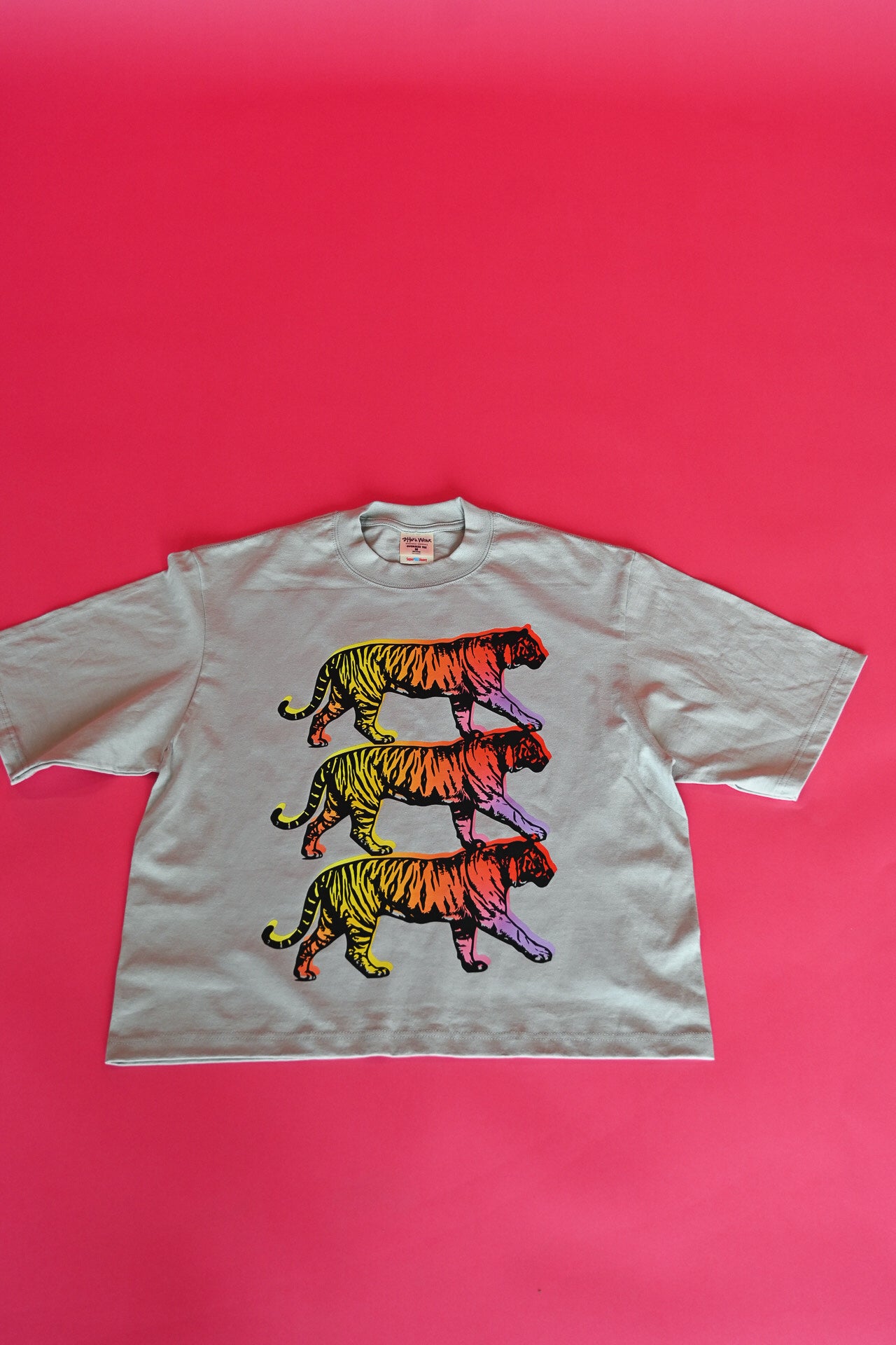 A light gray short-sleeve t-shirt featuring three stacked, colorful tiger illustrations in a rainbow gradient, laid flat against a vibrant pink background