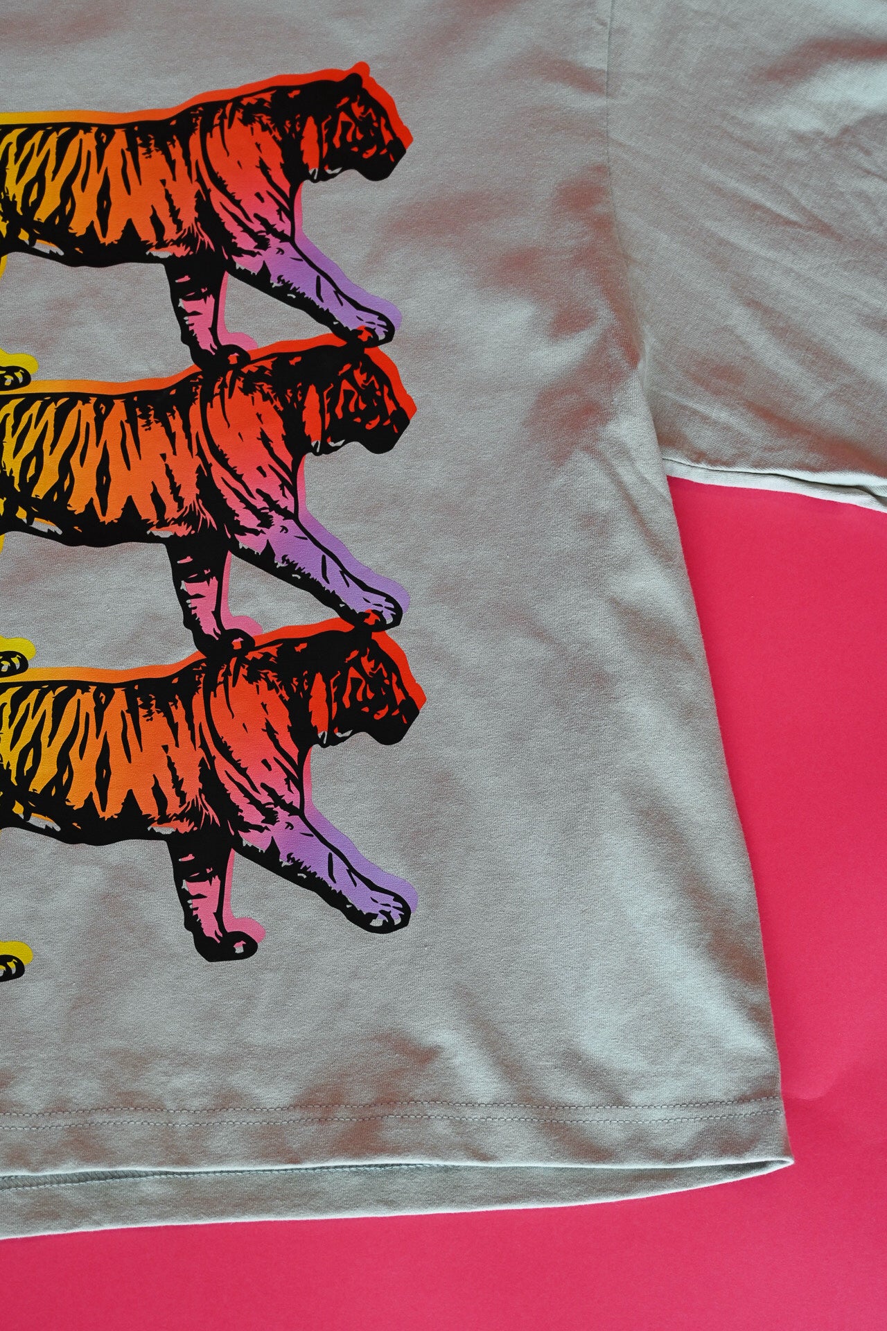 A close-up of a light gray t-shirt featuring a graphic print of three walking tigers in a vibrant gradient of orange, red, yellow, and purple. The t-shirt is laid flat on a bright pink background