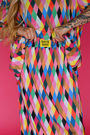 A close-up of a person wearing a bright multicolor diamond-pattern skirt with pleated texture and a yellow 'burnt toast' patch at the waist, posed against a solid pink background