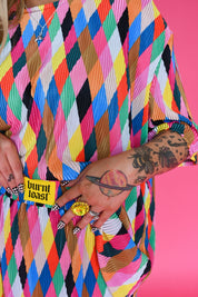 A close-up of a person wearing a bright multicolor pleated top with a repeating diagonal stripe/diamond pattern in pink, blue, yellow, orange, black, and white. The waistband has a yellow patch that reads 'burnt toast' against a solid pink background