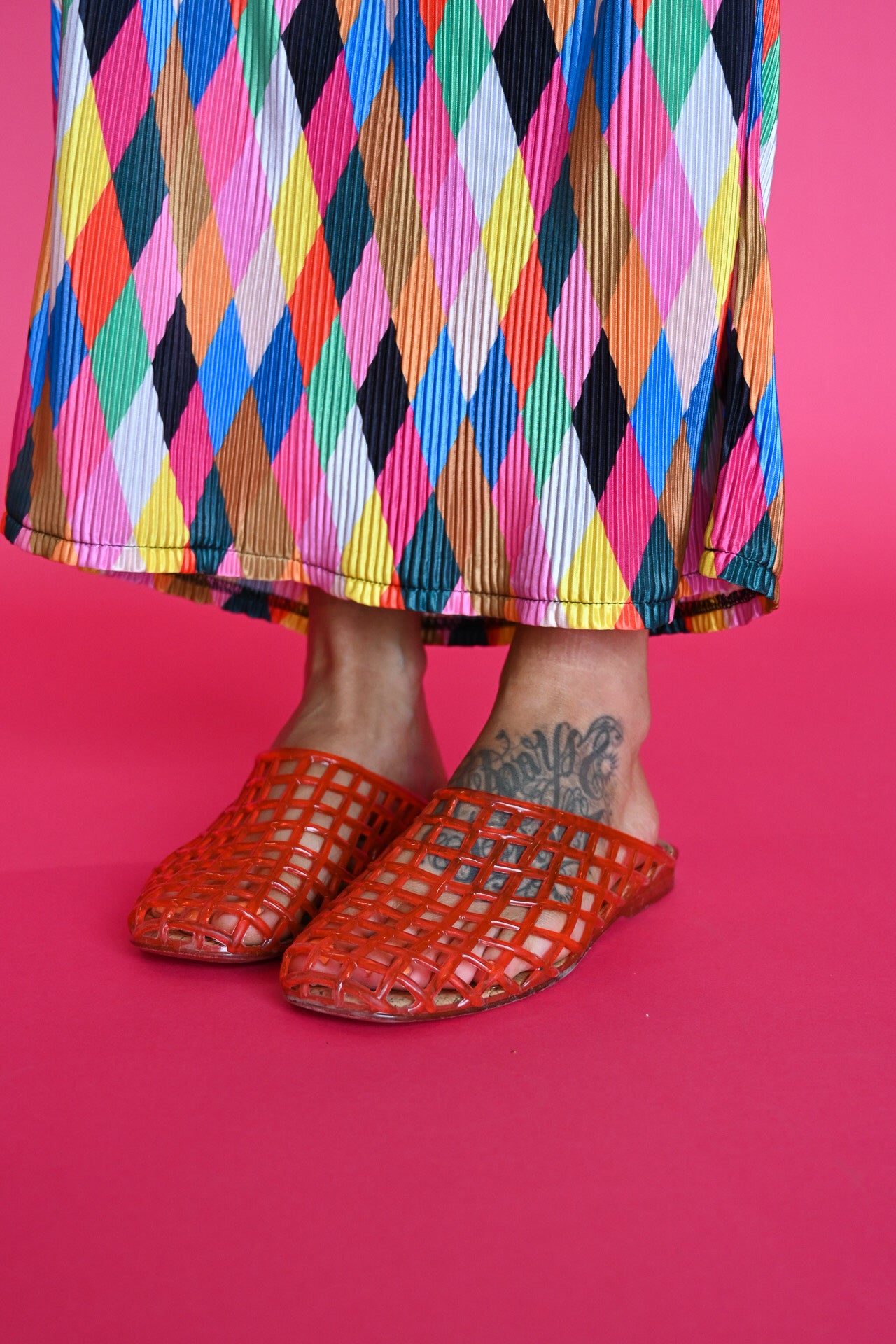 A person wearing bright orange jelly shoes and a colorful pleated skirt with a multicolor harlequin pattern, standing against a solid pink background