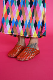 A person wearing bright orange jelly shoes and a colorful pleated skirt with a multicolor harlequin pattern, standing against a solid pink background