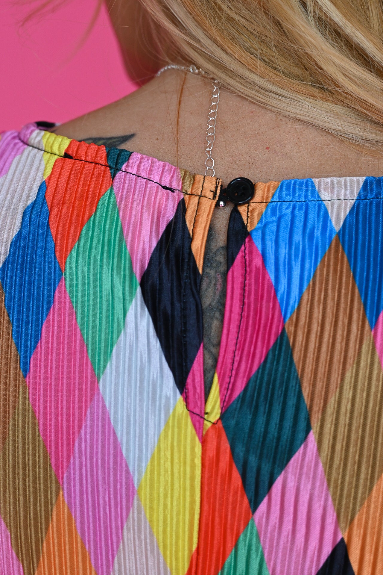 Close-up back view of a person wearing a brightly colored pleated top with a multicolor harlequin diamond pattern and a thin silver chain necklace