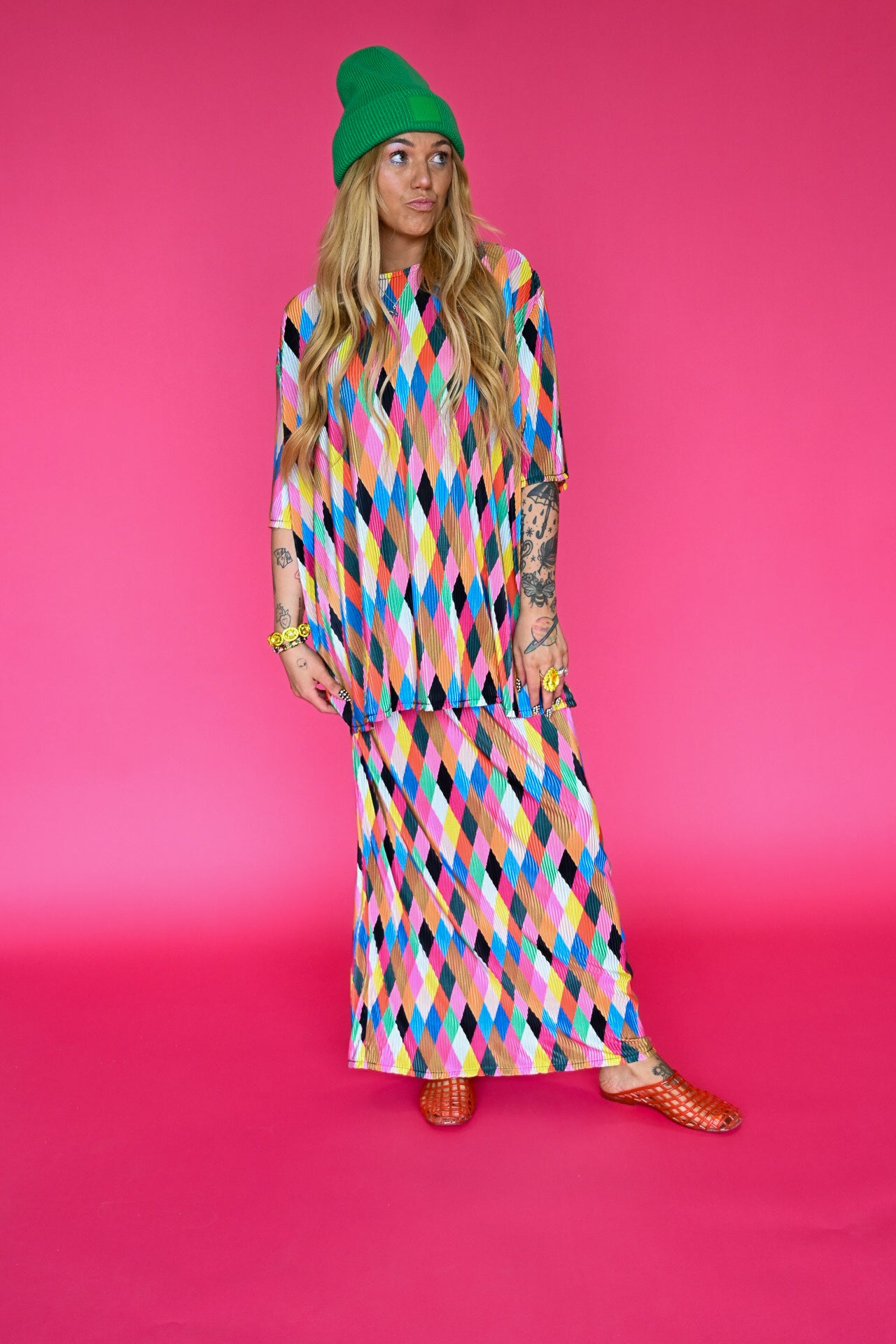 A model wearing a colorful geometric print maxi skirt and short sleeve top with a green beanie and gold flat sandals, standing against a solid hot pink studio background