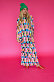 A model wearing a colorful geometric print maxi skirt and short sleeve top with a green beanie and gold flat sandals, standing against a solid hot pink studio background