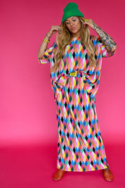 A woman modeling a vibrant, multicolored geometric print maxi skirt with plisse top with a yellow burnt toast tag on waistband, and green beanie, standing against a solid pink background