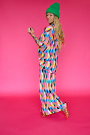 A woman modeling a colorful geometric print maxi skirt and short sleeve top, a green beanie, and orange jelly shoes against a solid pink studio background