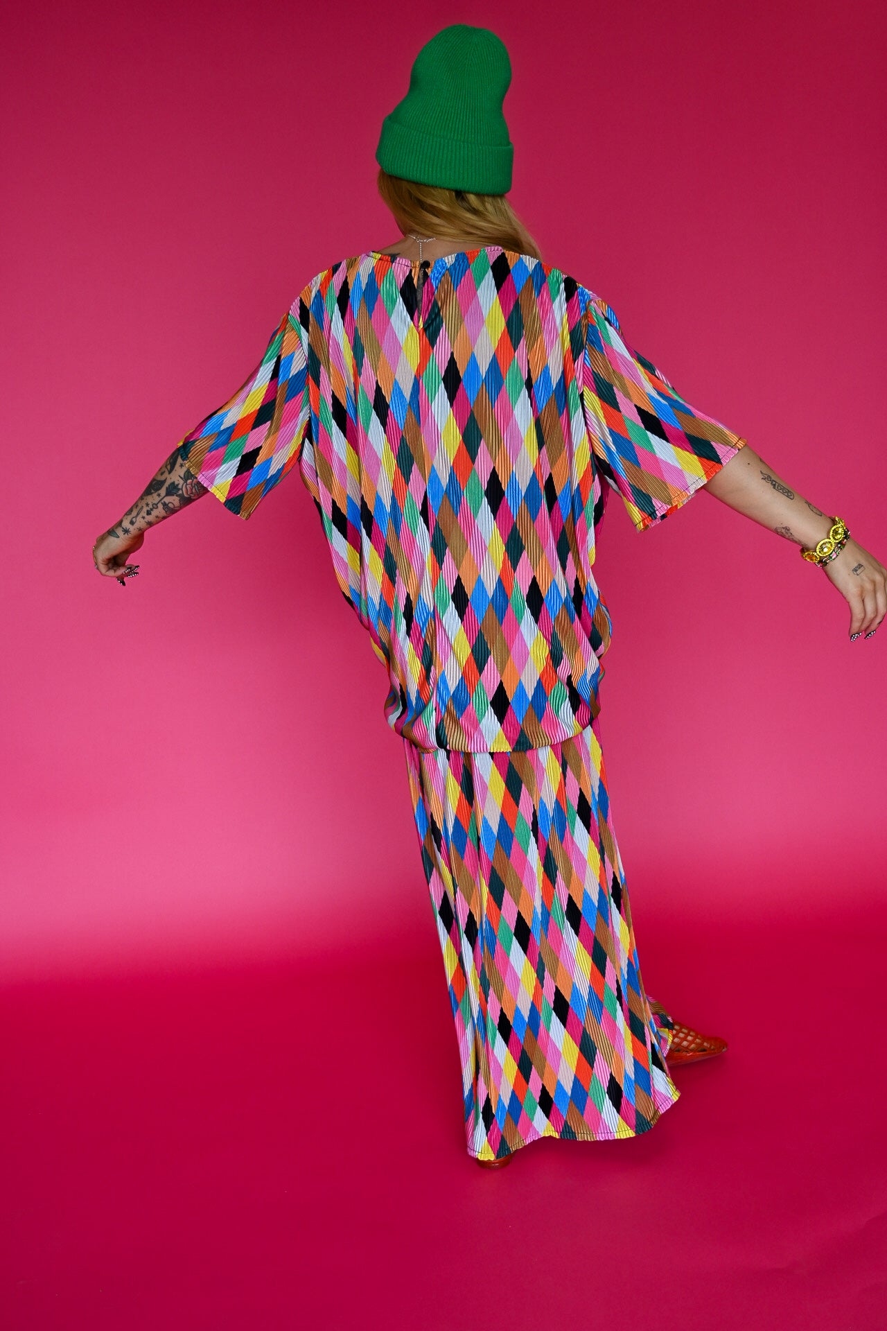 A person viewed from behind wearing a bright multicolor diamond-patterned matching set consisting of a loose short-sleeve top and maxi skirt, paired with a green beanie, against a solid hot pink studio background