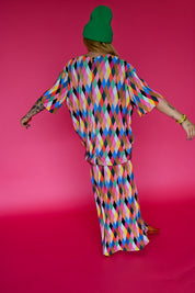 A person viewed from behind wearing a bright multicolor diamond-patterned matching set consisting of a loose short-sleeve top and maxi skirt, paired with a green beanie, against a solid hot pink studio background