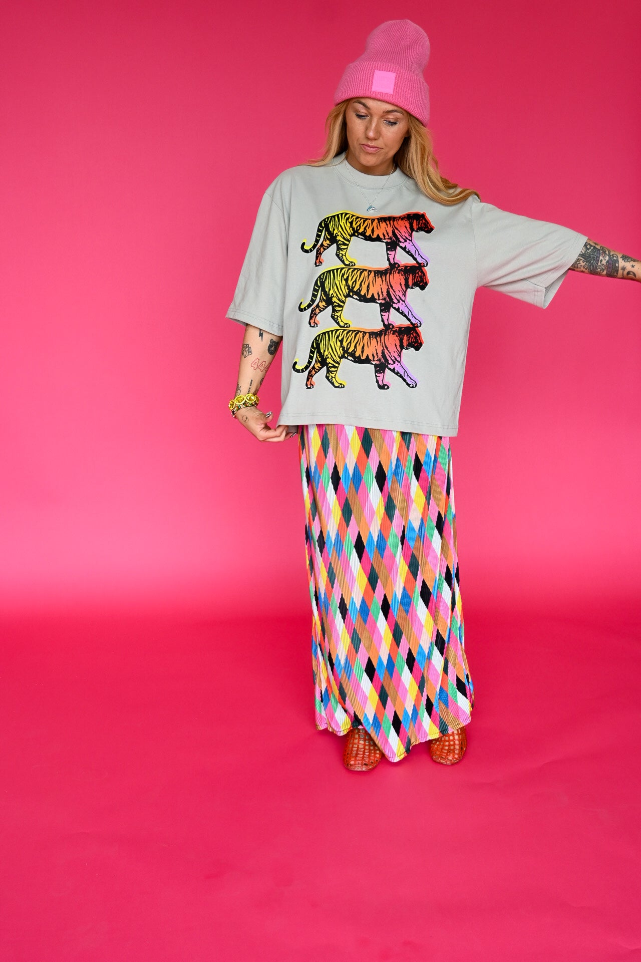 A woman modeling a light gray oversized t-shirt with a colorful stacked tiger graphic, paired with a long skirt featuring a vibrant multicolor diamond pattern, set against a solid hot pink studio background