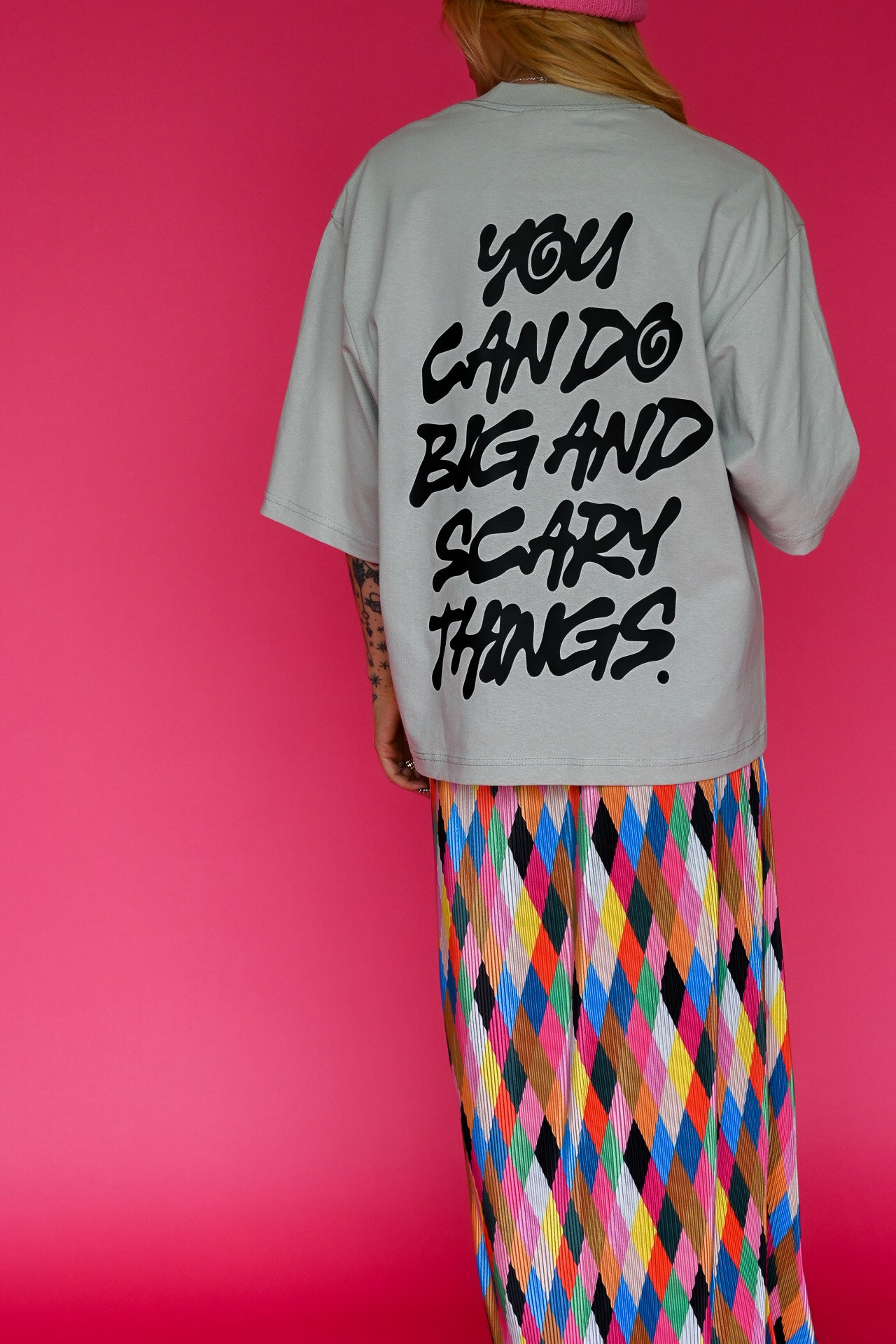 A person shown from the back wearing a light gray oversized T-shirt with bold black text reading 'YOU CAN DO BIG AND SCARY THINGS.' and a bright multicolor diamond-pattern skirt against a solid pink background