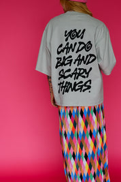 A person shown from the back wearing a light gray oversized T-shirt with bold black text reading 'YOU CAN DO BIG AND SCARY THINGS.' and a bright multicolor diamond-pattern skirt against a solid pink background