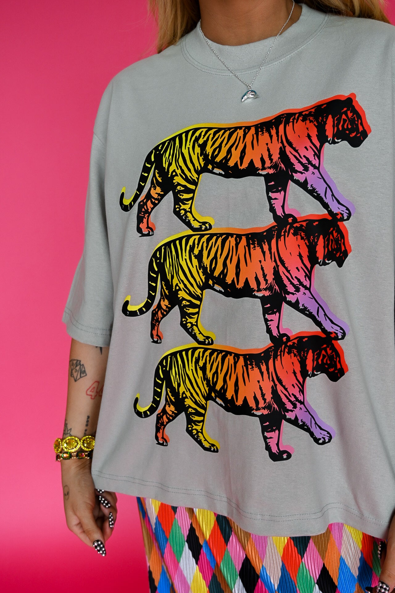 A person wearing an oversized light gray t-shirt with a graphic print of three walking tigers in a rainbow gradient, paired with a colorful argyle skirt, set against a solid pink background