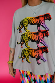 A person wearing an oversized light gray t-shirt with a graphic print of three walking tigers in a rainbow gradient, paired with a colorful argyle skirt, set against a solid pink background