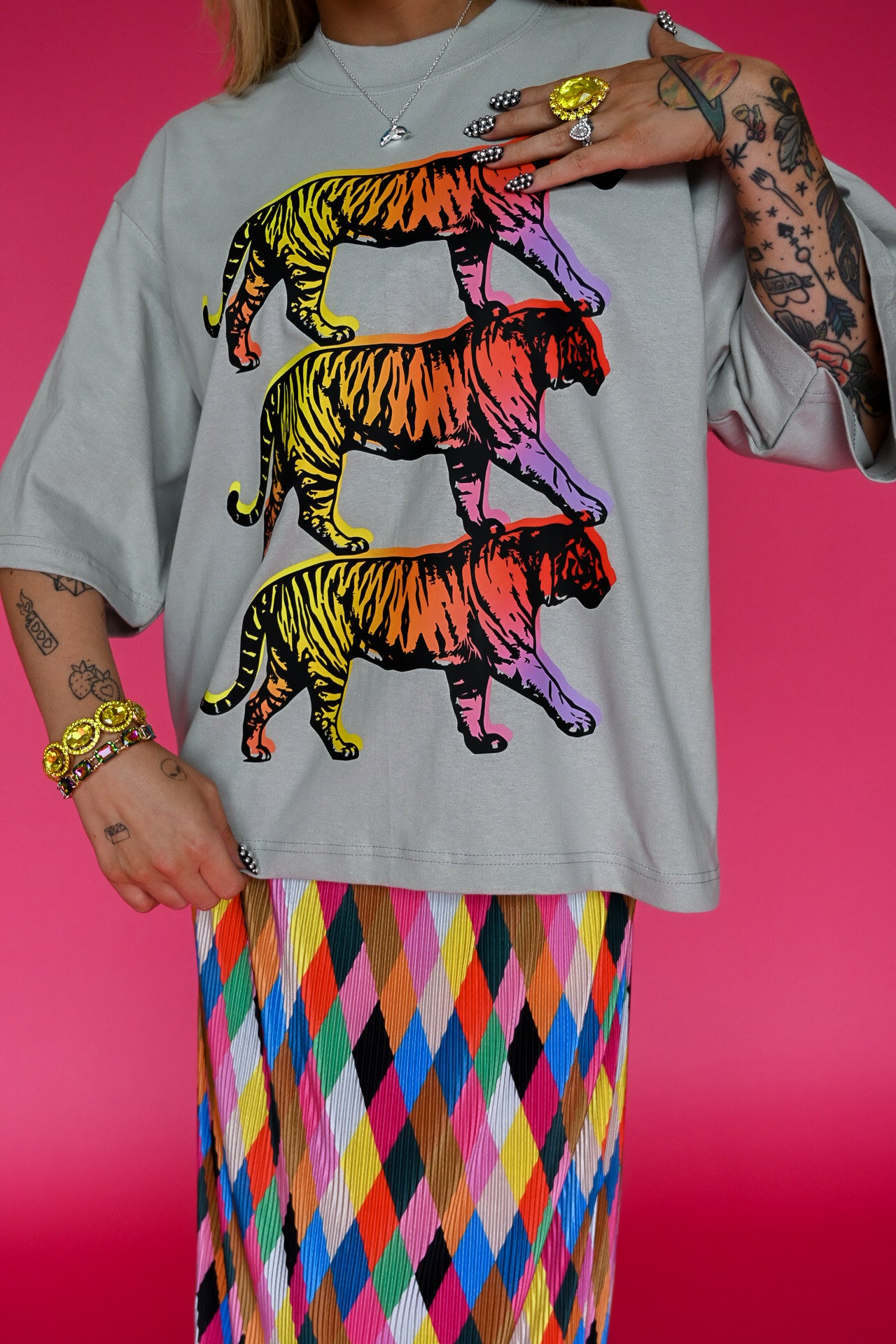 A person wearing an oversized light gray T-shirt with three stacked neon rainbow tiger graphics, paired with a colorful diamond-patterned skirt, against a bright pink background