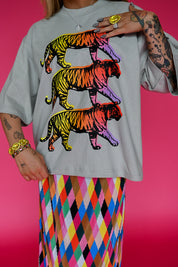 A person wearing an oversized light gray T-shirt with three stacked neon rainbow tiger graphics, paired with a colorful diamond-patterned skirt, against a bright pink background