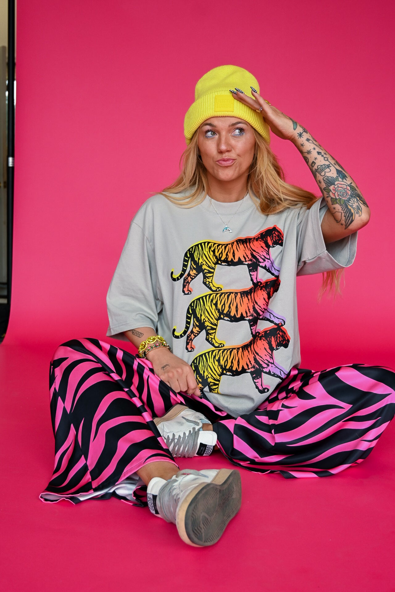 A woman sits cross-legged on a bright pink studio background, wearing a light gray oversized T-shirt with three colorful tiger graphics in orange, yellow, and green, a bright yellow beanie, and pink-and-black zebra-print pants