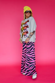 A model stands against a vibrant pink studio background wearing a light gray oversized t-shirt with a colorful tiger graphic, wide-leg pink and black zebra-print pants, a yellow beanie, and sunglasses