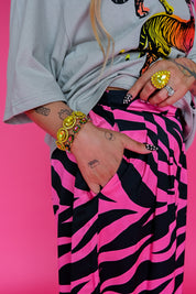 A close-up lifestyle shot of a person wearing a light gray t-shirt with a colorful graphic and hot pink and black zebra-print pants, accessorized with gold bracelets and rings, set against a solid hot pink background