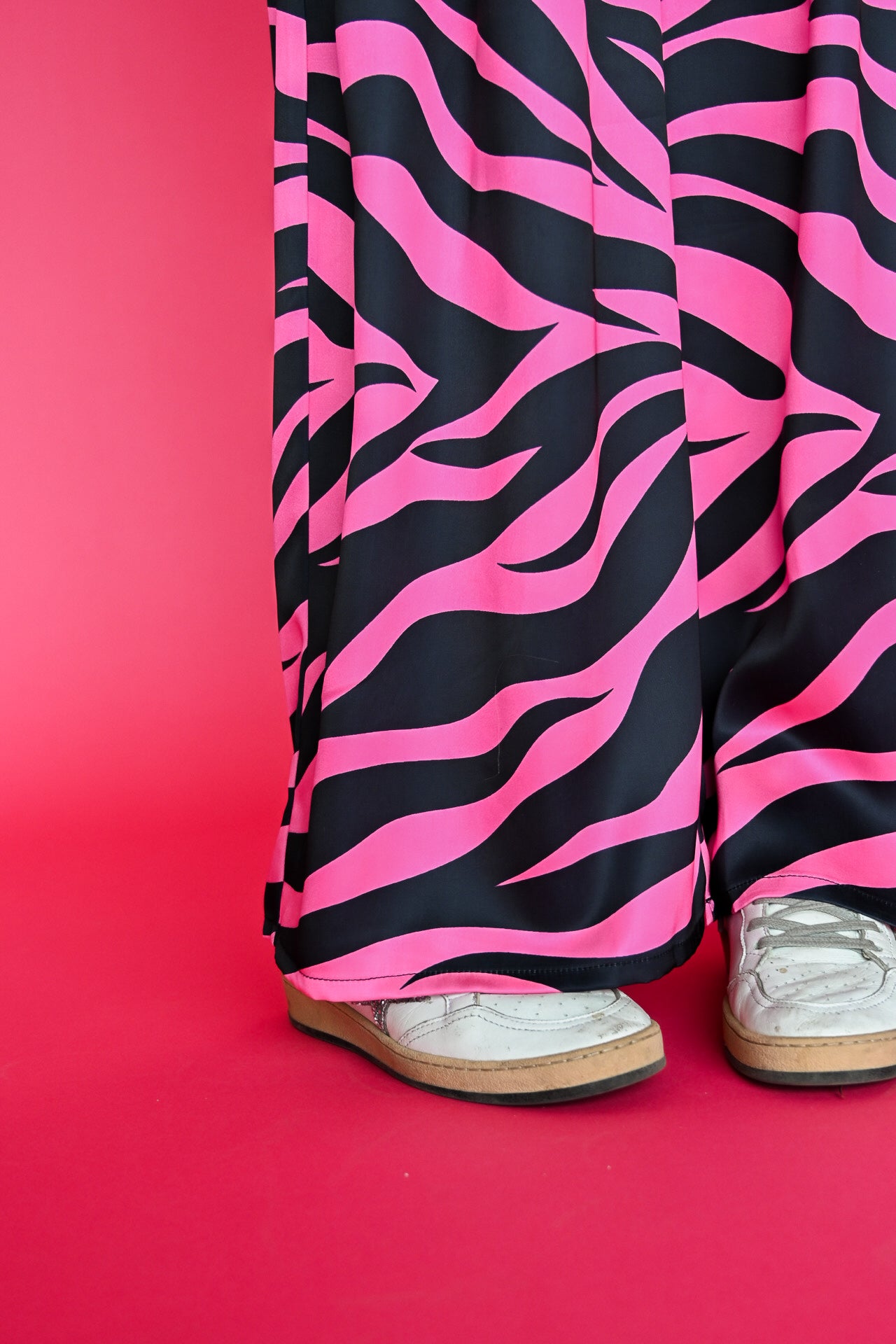 Pink and black zebra-print wide-leg pants worn with white sneakers, shown from the knees down against a solid pink background