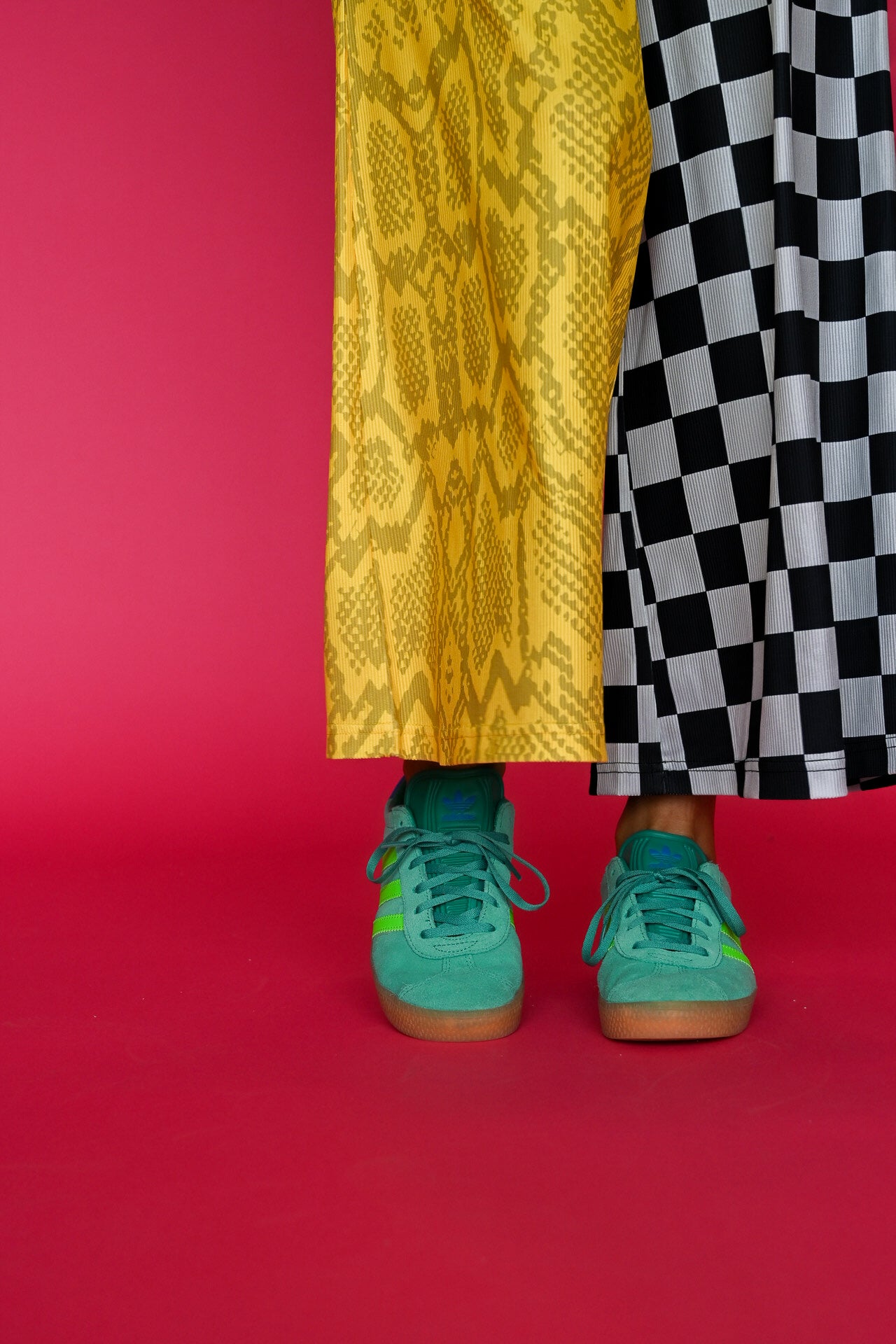 Close up of half and half pants with yellow snakeskin and black and white checkered design, and green adidas shoes, on a pink background