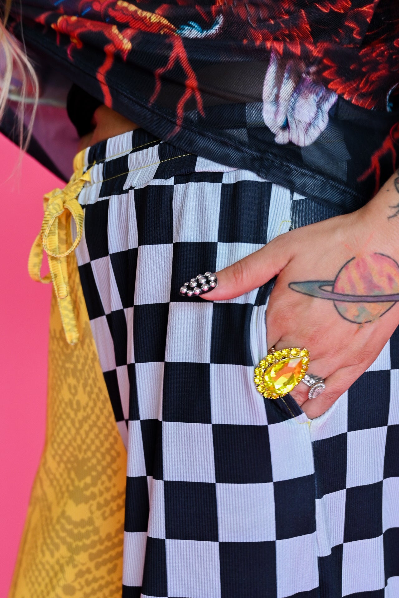 Close-up of a person wearing black and white checkered pants with a yellow drawstring, a black graphic top, and multiple rings on their hand