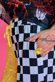 Close-up of a person wearing black and white checkered pants with a yellow drawstring, a black graphic top, and multiple rings on their hand