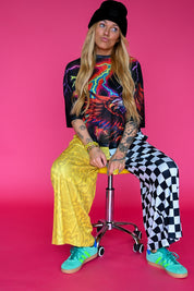 A woman sits on a yellow stool against a solid pink studio background, wearing a black beanie, a colorful neon graphic t-shirt, a shiny gold pleated pants, black-and-white checkered wide-leg pants, and bright turquoise sneakers
