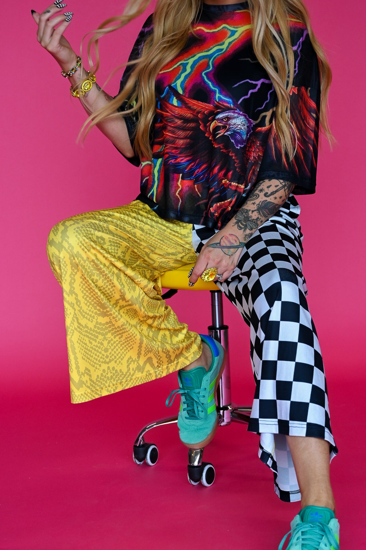 A model wearing a vibrant graphic mesh top with a colorful eagle design, yellow snakeskin wide-leg pants, and a black-and-white checkered leg, sitting on a rolling stool against a solid pink background