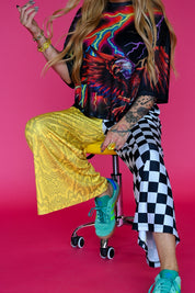 A model wearing a vibrant graphic mesh top with a colorful eagle design, yellow snakeskin wide-leg pants, and a black-and-white checkered leg, sitting on a rolling stool against a solid pink background