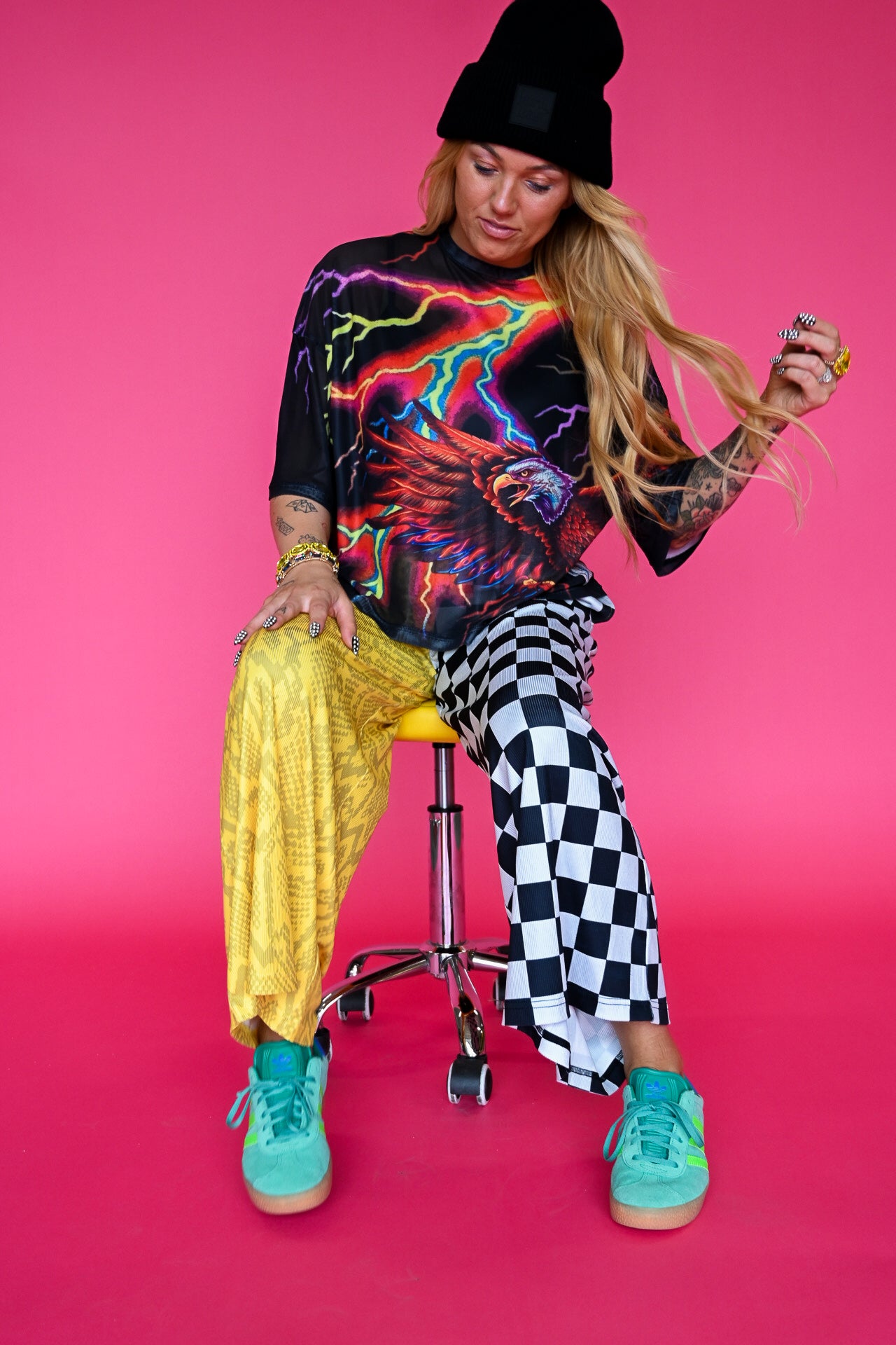 A woman sits on a yellow stool against a bright pink studio background, wearing a black beanie, a black graphic t-shirt with neon lightning and an eagle design, yellow snakeskin-print pants, black-and-white checkered pants, and turquoise sneakers