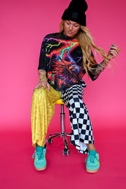 A woman sits on a yellow stool against a bright pink studio background, wearing a black beanie, a black graphic t-shirt with neon lightning and an eagle design, yellow snakeskin-print pants, black-and-white checkered pants, and turquoise sneakers