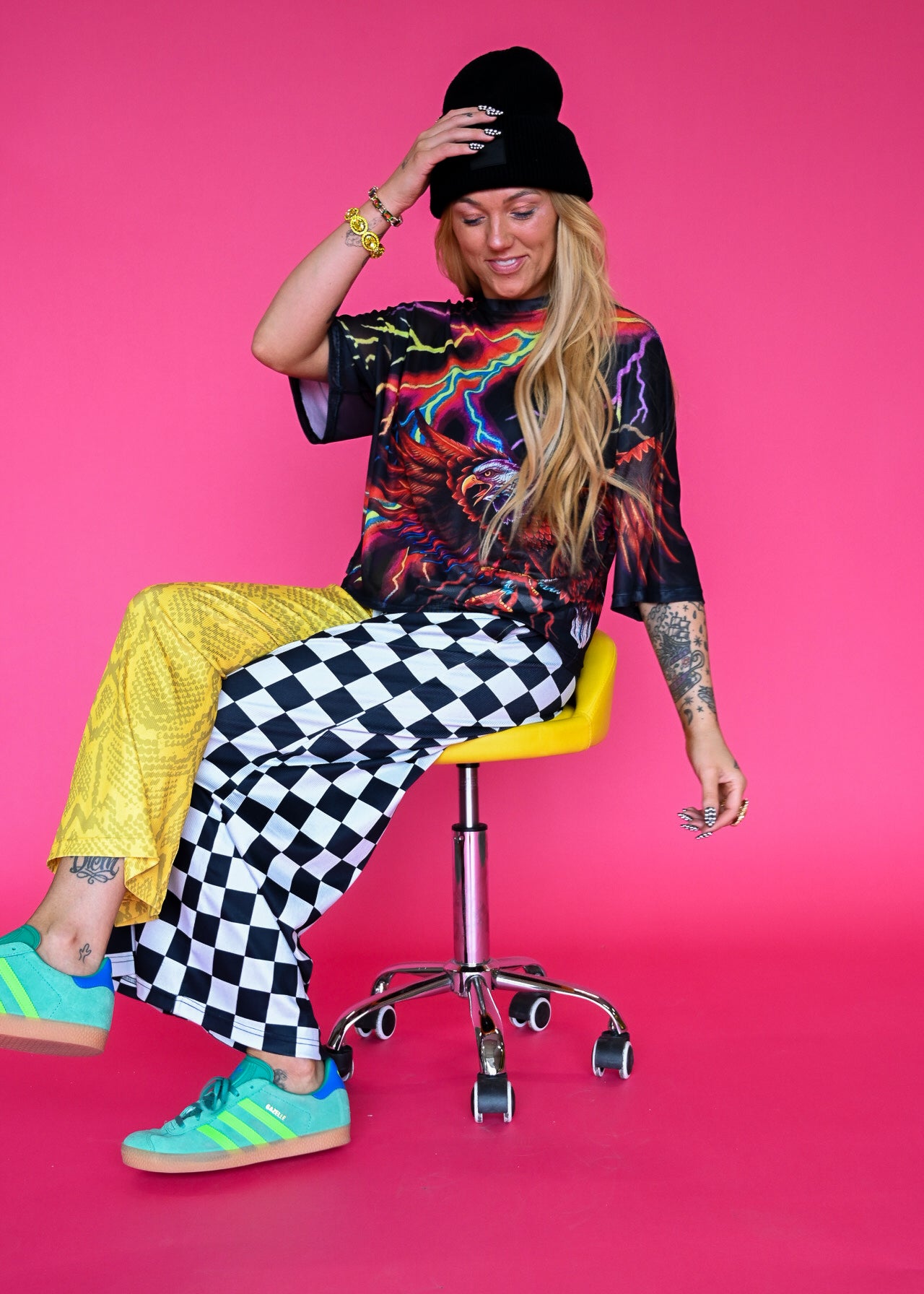 A woman sits on a yellow and chrome swivel stool against a solid hot pink studio background. She is wearing a black beanie, a black graphic t-shirt, bright yellow pants, black and white checkered wide-leg pants, and teal sneakers with yellow stripes