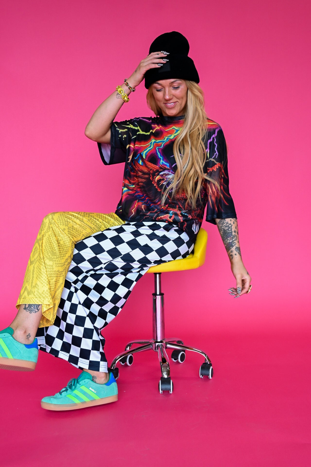 A woman sits on a yellow and chrome swivel stool against a solid hot pink studio background. She is wearing a black beanie, a black graphic t-shirt, bright yellow pants, black and white checkered wide-leg pants, and teal sneakers with yellow stripes