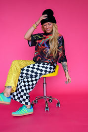 A woman sits on a yellow and chrome swivel stool against a solid hot pink studio background. She is wearing a black beanie, a black graphic t-shirt, bright yellow pants, black and white checkered wide-leg pants, and teal sneakers with yellow stripes