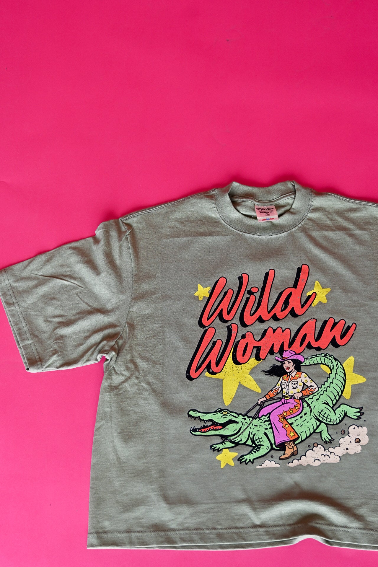 A vintage-style short-sleeve graphic t-shirt laid flat on a vibrant pink background. The t-shirt is a muted olive green with a colorful graphic of a woman riding a green alligator, with the text 'Wild Woman' in red and yellow
