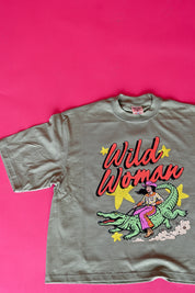 A vintage-style short-sleeve graphic t-shirt laid flat on a vibrant pink background. The t-shirt is a muted olive green with a colorful graphic of a woman riding a green alligator, with the text 'Wild Woman' in red and yellow